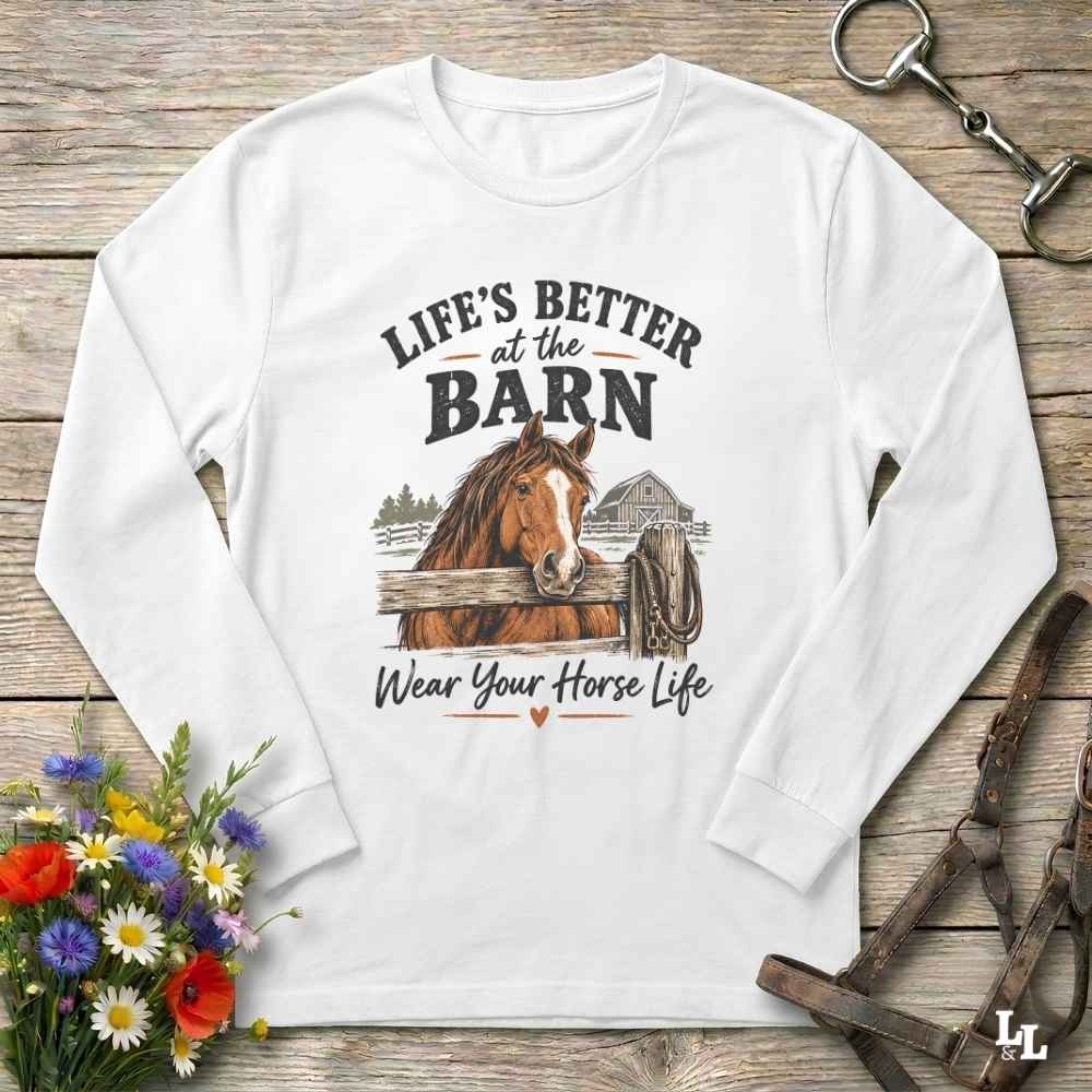 A white long-sleeve crewneck sweatshirt with a rustic equestrian graphic on the front. The graphic features a brown horse, a barn, and the text 'LIFE'S BETTER at the BARN Wear Your Horse Life' in black and brown.