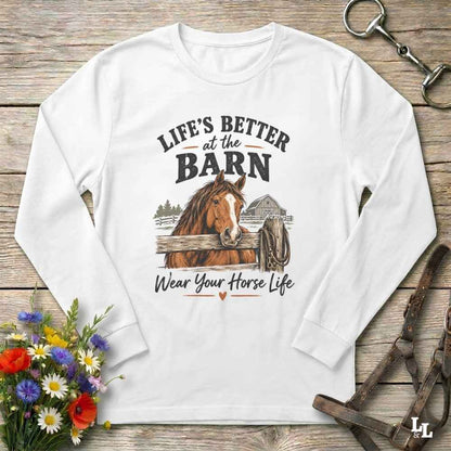 A white long-sleeve crewneck sweatshirt with a rustic equestrian graphic on the front. The graphic features a brown horse, a barn, and the text 'LIFE'S BETTER at the BARN Wear Your Horse Life' in black and brown.