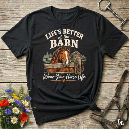 A black graphic t-shirt laid flat on a rustic wooden surface, featuring a brown horse illustration and the text 'LIFE'S BETTER at the BARN Wear Your Horse Life'. The shirt is styled with equestrian accessories and wildflowers.