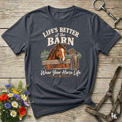 A dark heather gray graphic t-shirt featuring a brown horse and the text 'LIFE'S BETTER at the BARN Wear Your Horse Life' in a styled flat-lay on a rustic wooden background with flowers and horse tack.