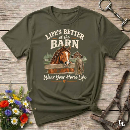 An olive green graphic t-shirt laid flat on a rustic wooden background, styled with equestrian accessories and a small bouquet of wildflowers. The shirt features a graphic of a brown horse and the text 'LIFE'S BETTER at the BARN Wear Your Horse Life'.