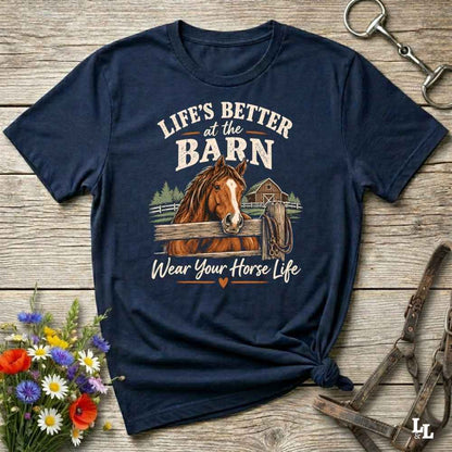 A navy blue graphic t-shirt featuring a brown horse and a barn illustration with the text 'LIFE'S BETTER at the BARN Wear Your Horse Life'. The t-shirt is styled in a flat-lay on a rustic wooden background with equestrian accessories and flowers.