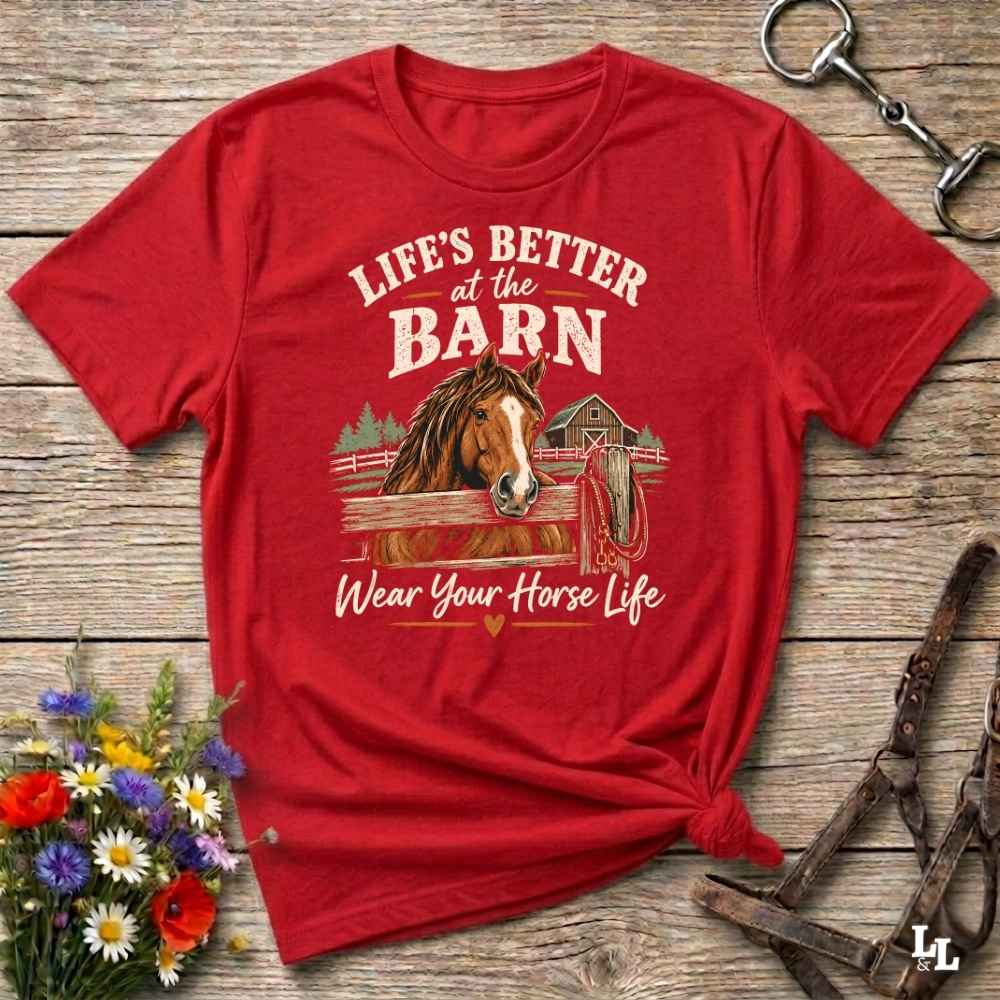 A red short-sleeve graphic T-shirt laid flat on a rustic wooden surface, styled with flowers, a horse bit, and a bridle. The shirt features a large front print with the words “LIFE'S BETTER at the BARN” and “Wear Your Horse Life” around an illustration of a horse at a barn.
