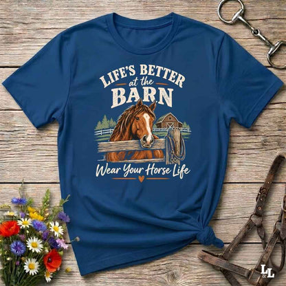 A navy blue graphic t-shirt laid flat on a rustic wooden surface, styled with a bouquet of wildflowers, a horse bit, and a leather halter. The shirt features a graphic of a brown horse behind a fence with the text 'LIFE'S BETTER at the BARN Wear Your Horse Life'.