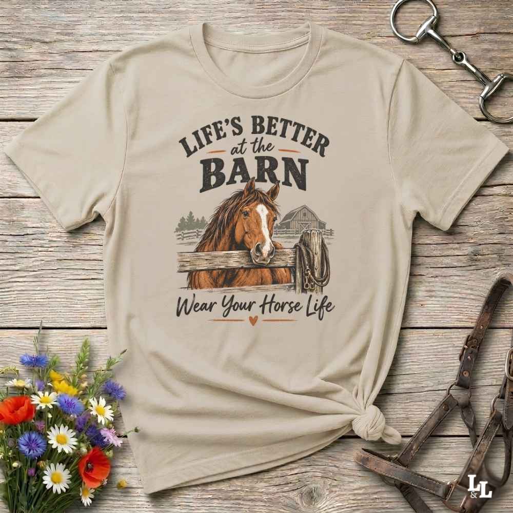 A beige graphic t-shirt featuring a brown horse and the text 'LIFE'S BETTER at the BARN Wear Your Horse Life' printed on the front, styled in a flat-lay on a rustic wooden background with flowers and horse tack.