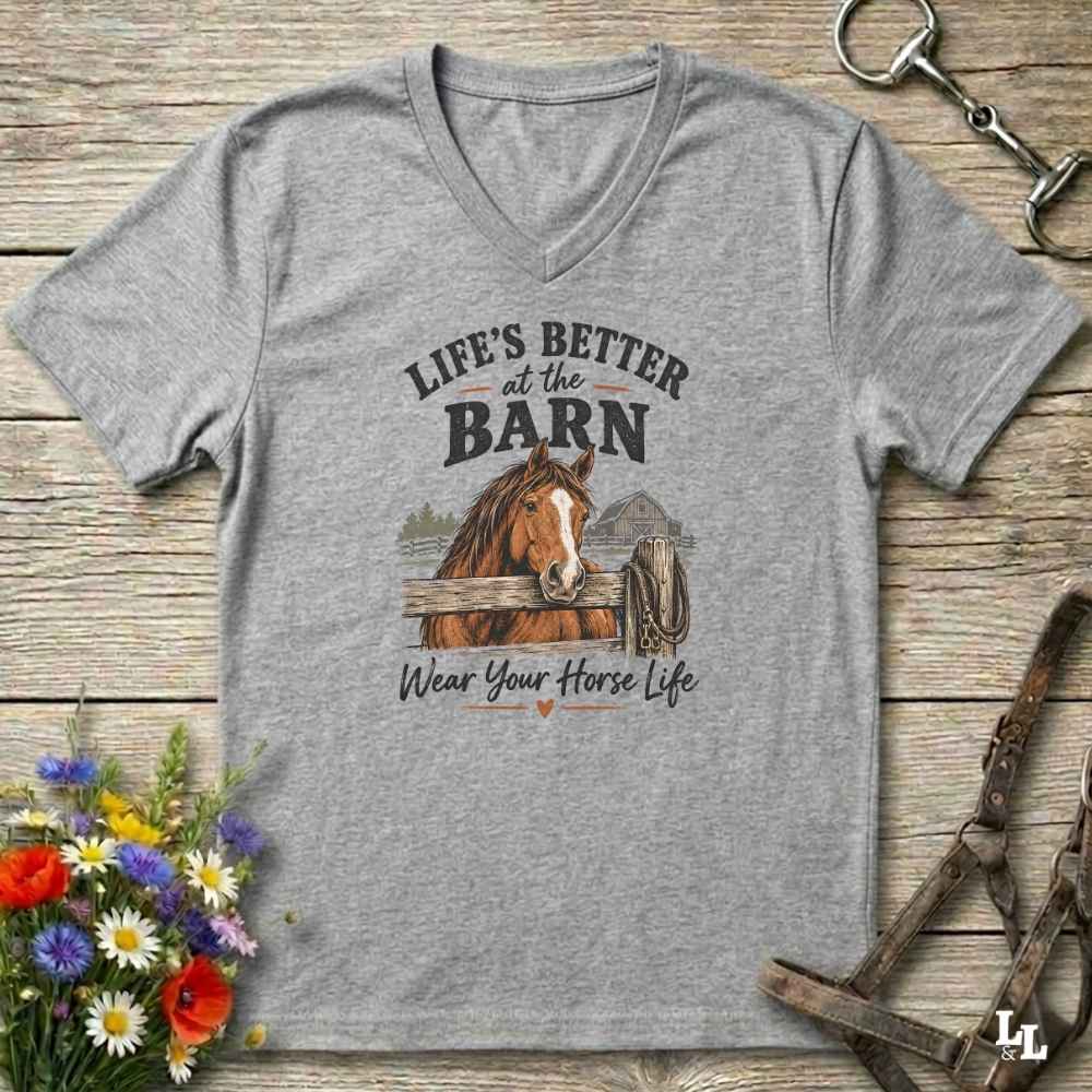 A heather gray V-neck t-shirt with a graphic of a brown horse and the text 'LIFE'S BETTER at the BARN' and 'Wear Your Horse Life', displayed on a rustic wooden background with equestrian accessories and flowers.