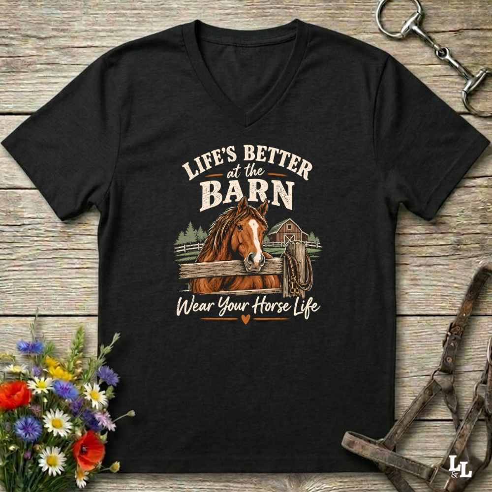 A black V-neck t-shirt with a graphic print of a brown horse and the text 'LIFE'S BETTER at the BARN Wear Your Horse Life', displayed on a rustic wooden background with equestrian accessories and flowers.