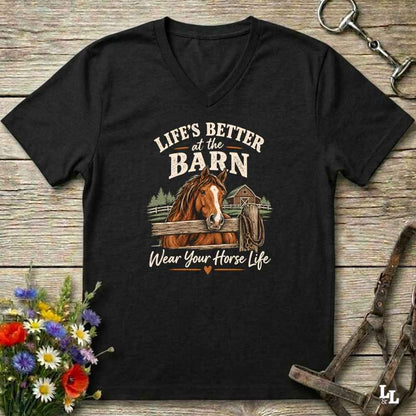 A black V-neck t-shirt with a graphic print of a brown horse and the text 'LIFE'S BETTER at the BARN Wear Your Horse Life', displayed on a rustic wooden background with equestrian accessories and flowers.