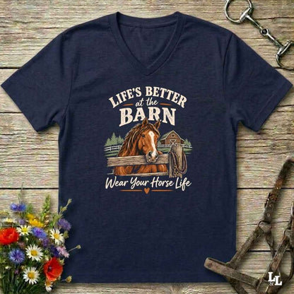 A navy blue V-neck t-shirt with a graphic print of a brown horse and the text 'LIFE'S BETTER at the BARN Wear Your Horse Life', displayed on a rustic wooden background with equestrian accessories and flowers.