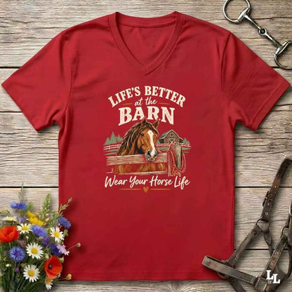 A red V-neck t-shirt with a graphic print of a horse and the text 'LIFE'S BETTER at the BARN Wear Your Horse Life', displayed on a rustic wooden background with equestrian accessories and flowers.