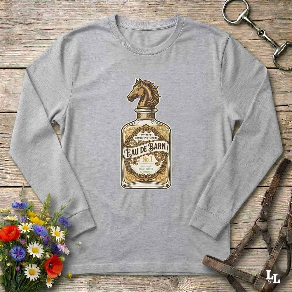 A heather gray long-sleeve t-shirt with a vintage-style graphic of a perfume bottle labeled 'Eau de Barn' featuring a horse head. The shirt is displayed flat on a rustic wooden background with equestrian and floral props.