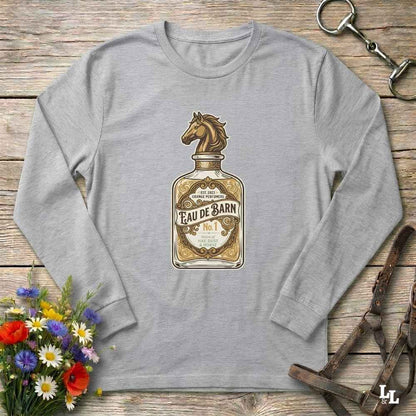 A heather gray long-sleeve t-shirt with a vintage-style graphic of a perfume bottle labeled 'Eau de Barn' featuring a horse head. The shirt is displayed flat on a rustic wooden background with equestrian and floral props.