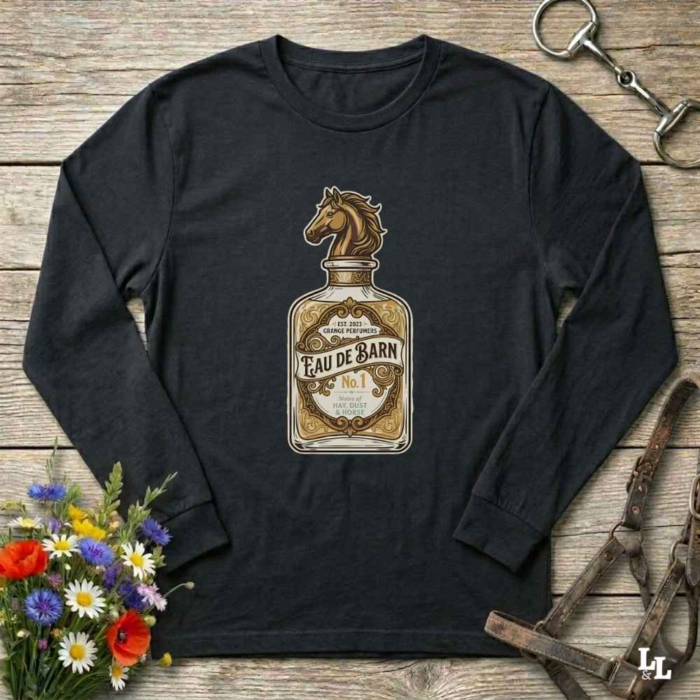 A black long-sleeve t-shirt with a vintage-style 'Eau de Barn' whiskey bottle graphic featuring a horse head. The shirt is displayed in a flat-lay arrangement on a rustic wooden background, surrounded by equestrian leather tack and a small bouquet of wildflowers.