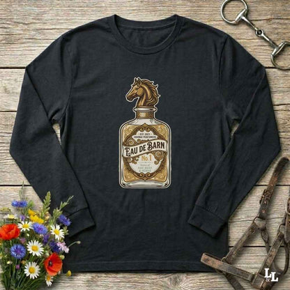 A black long-sleeve t-shirt with a vintage-style 'Eau de Barn' whiskey bottle graphic featuring a horse head. The shirt is displayed in a flat-lay arrangement on a rustic wooden background, surrounded by equestrian leather tack and a small bouquet of wildflowers.