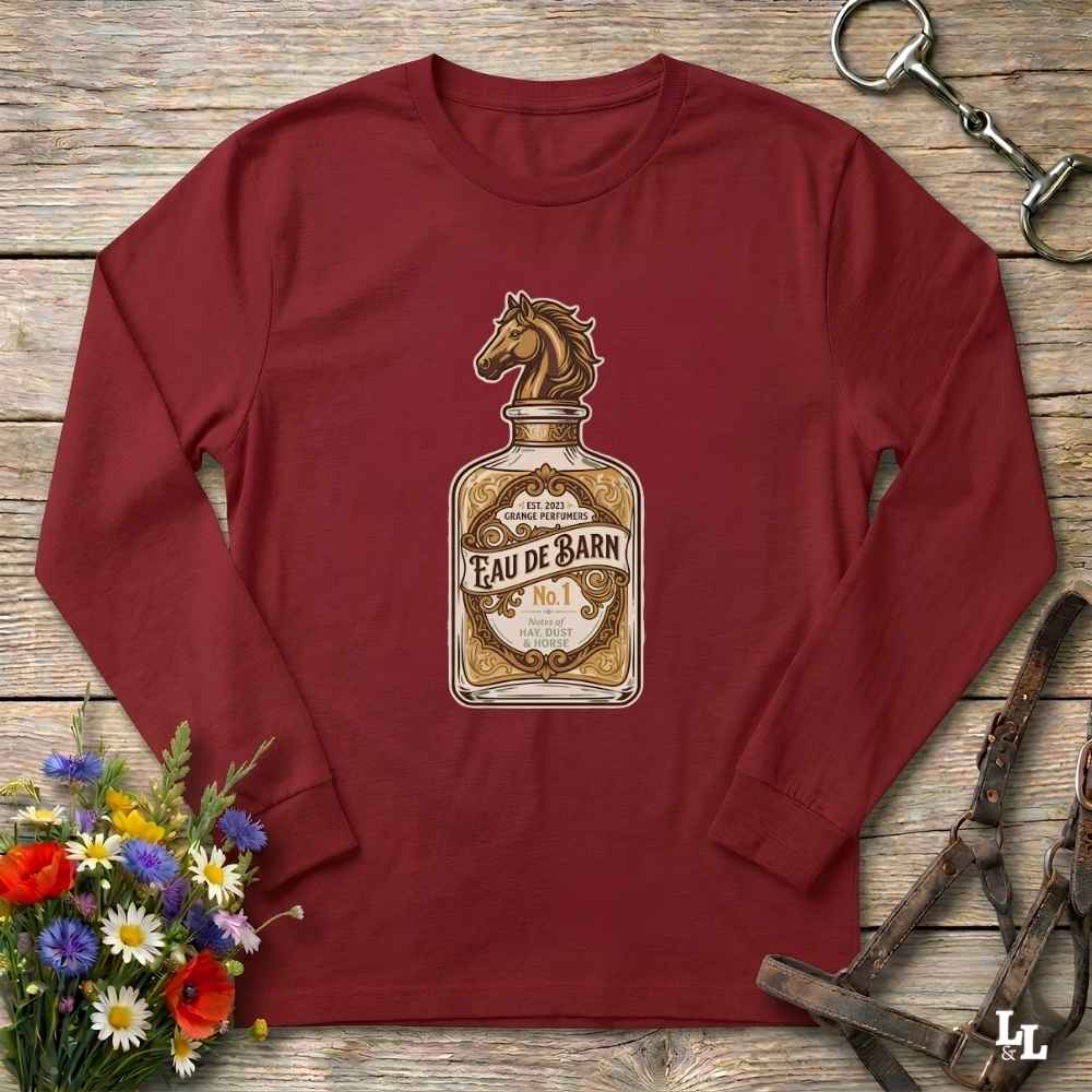 A long-sleeve crewneck t-shirt in maroon red, featuring a graphic print of a vintage perfume bottle labeled 'Eau de Barn' with a horse head on the cap. The shirt is displayed flat on a rustic wooden background, styled with equestrian tack and a small bouquet of wildflowers.