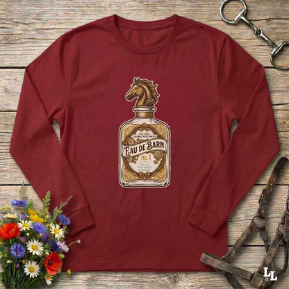 A long-sleeve crewneck t-shirt in maroon red, featuring a graphic print of a vintage perfume bottle labeled 'Eau de Barn' with a horse head on the cap. The shirt is displayed flat on a rustic wooden background, styled with equestrian tack and a small bouquet of wildflowers.