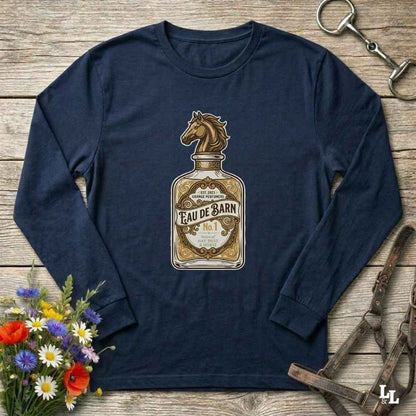 A navy blue long-sleeve t-shirt with a graphic of a vintage Eau de Barn bottle and horse head, displayed flat on a rustic wooden background with equestrian and floral props.