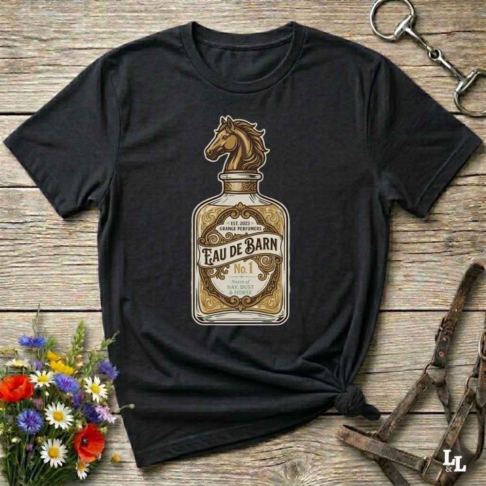 A black short-sleeve graphic T-shirt laid flat on a rustic wooden surface, featuring a large vintage-style bottle label design with a horse head and the text 'Fau De Barn No. 1.