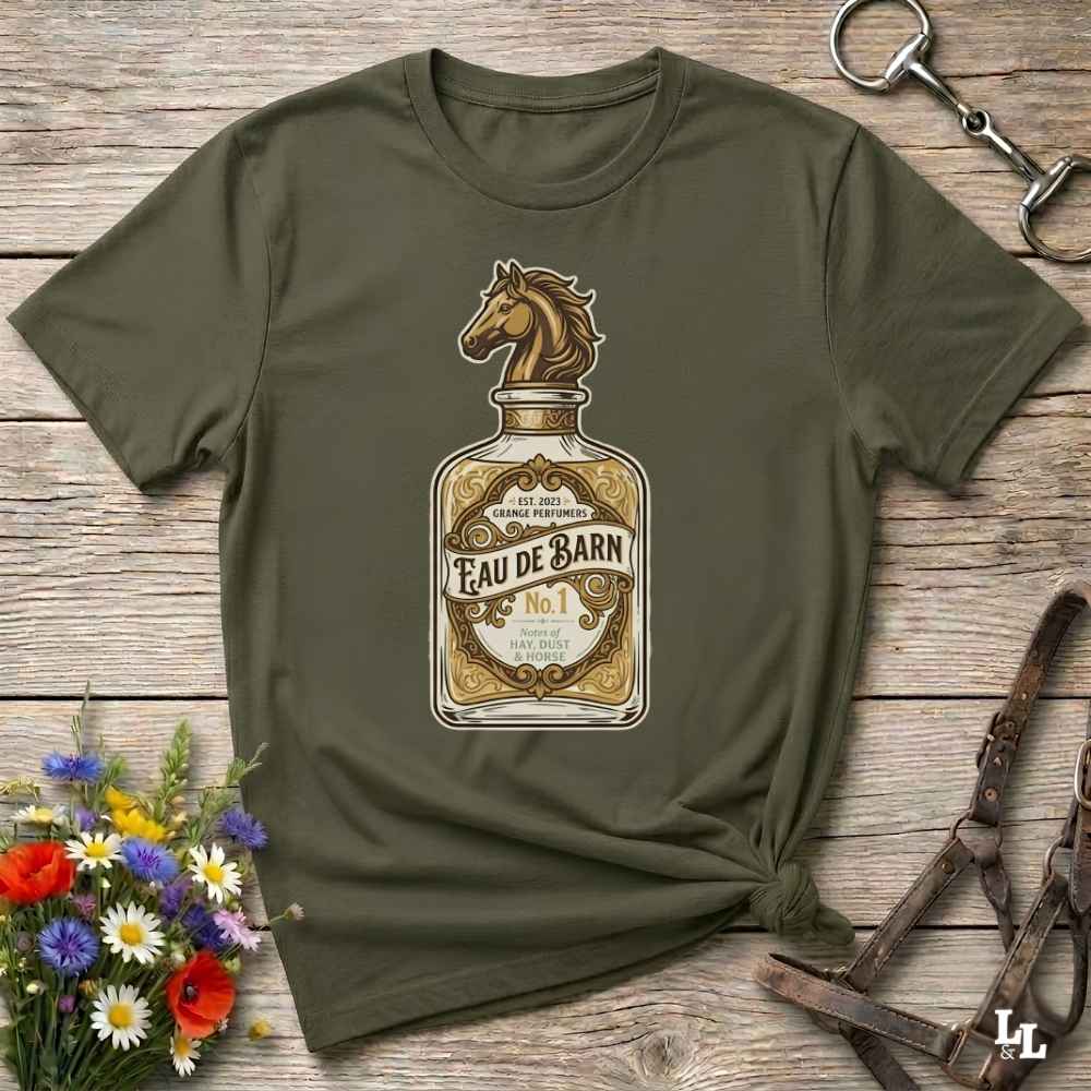 An olive green short-sleeve t-shirt with a graphic of a horse bottle label reading 'Feau de Barn No. 1', styled in a flat-lay on a rustic wooden background with equestrian accessories and flowers.