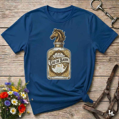 A navy blue short-sleeve t-shirt with a graphic of a horse-shaped perfume bottle labeled 'Feau de Barn No. 1', styled flat on a rustic wooden background with equestrian accessories and flowers.