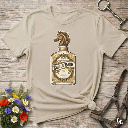 A beige short-sleeve t-shirt with a graphic of a horse-shaped bottle labeled 'Feau de Barn No. 1', styled flat on a rustic wooden background with equestrian accessories and a small flower arrangement.