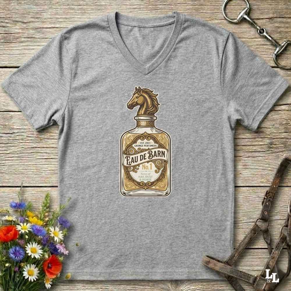 A heather gray V-neck t-shirt laid flat on a rustic wooden surface, featuring a vintage-style graphic of an eau de barn bottle with a horse head on the label. The shirt is styled with equestrian accessories (a horse bit and bridle) and a small bouquet of colorful wildflowers.