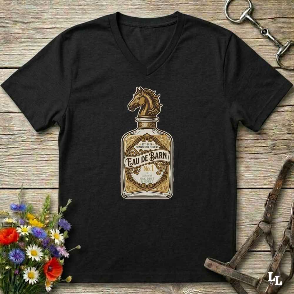 A black V-neck t-shirt laid flat on a rustic wooden surface, featuring a vintage-style graphic of an Eau de Barn bottle with a horse head on the neck. The shirt is styled with equestrian accessories, including a horse bit in the top right and a bridle strap in the bottom right, with a small bouquet of wildflowers in the bottom left.