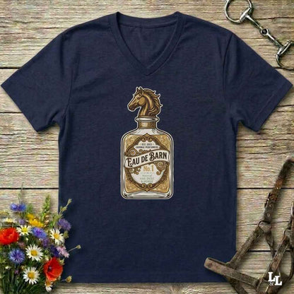 A navy blue v-neck t-shirt laid flat on a rustic wooden surface, featuring a vintage-style graphic of an Eau de Barn bottle with a horse head illustration. The shirt is styled with equestrian-themed props including a halter, tack, and a small bouquet of wildflowers.