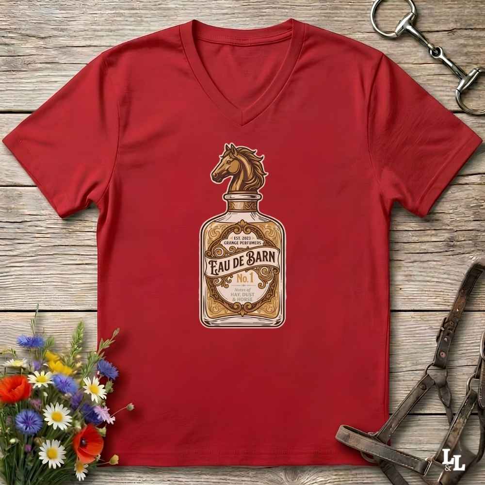 A red V-neck t-shirt laid flat on a rustic wooden surface, featuring a graphic of an Eau de Barn bottle with a horse head illustration. The shirt is styled with equestrian tack in the background and a small bouquet of colorful wildflowers in the lower-left corner.