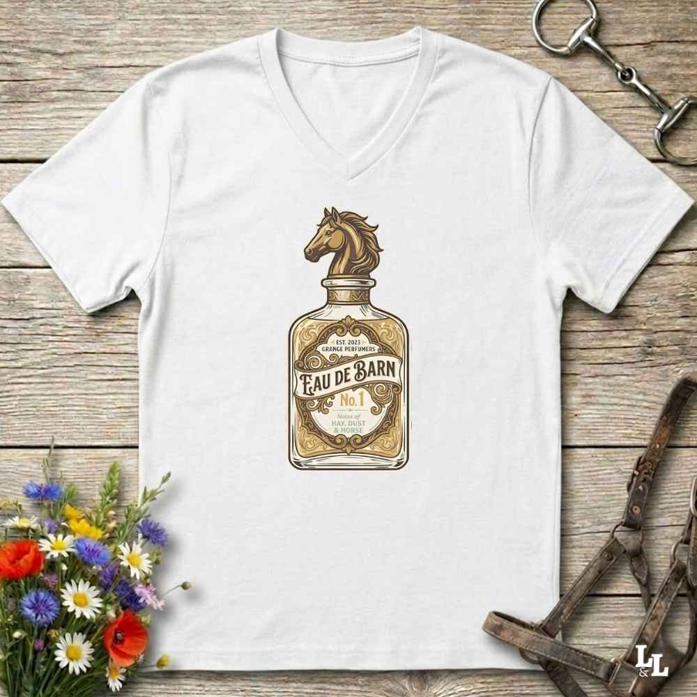 A white V-neck t-shirt laid flat on a rustic wooden surface, featuring a vintage-style graphic of an 'Eau de Barn' bottle with a horse head on the label. The shirt is styled with equestrian-themed props, including a horse bit in the top right and a leather bridle strap in the bottom right, with a small bouquet of colorful flowers in the bottom left.