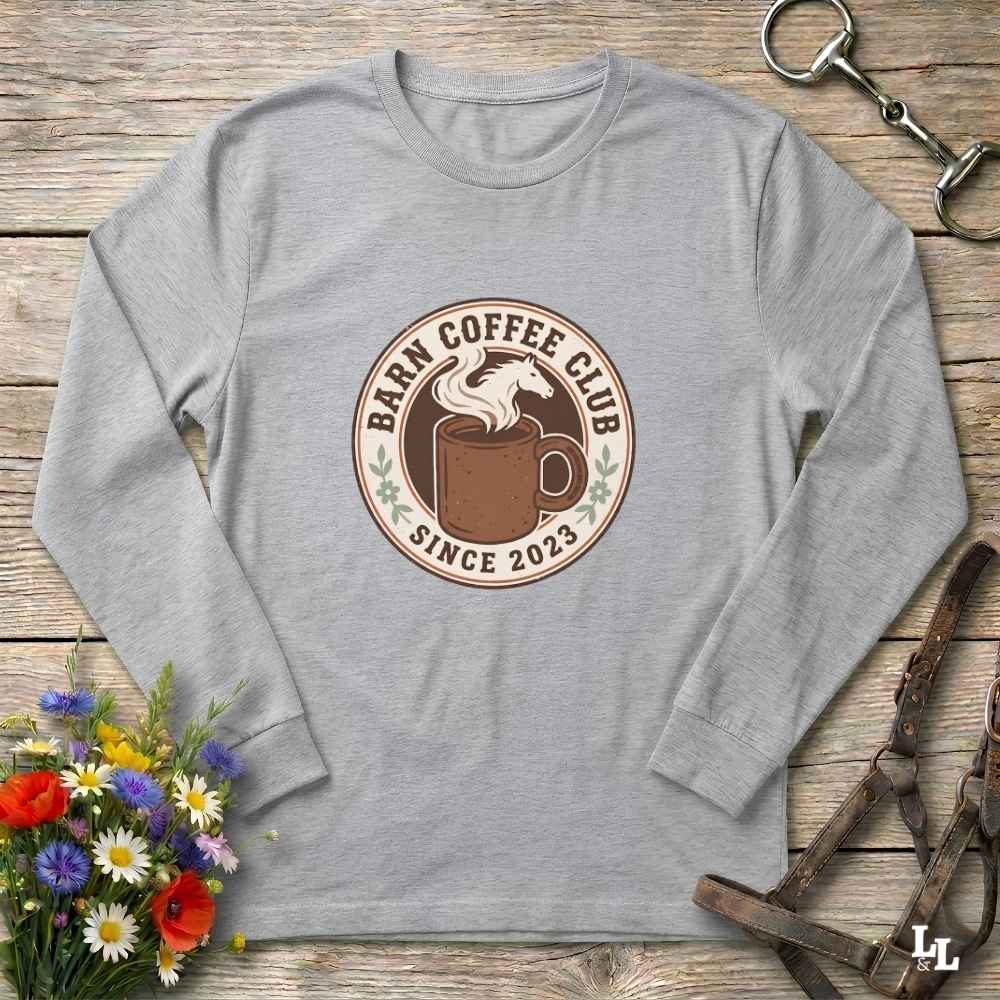 A heather gray long-sleeve t-shirt with a circular 'Barm Coffee Club' graphic on the chest, styled in a flat-lay on a rustic wooden background with flowers and horse tack.