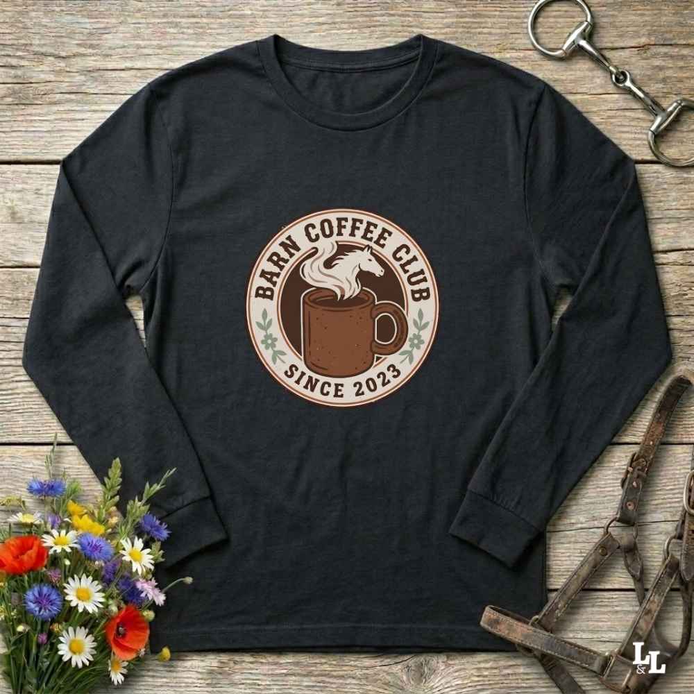 A black long-sleeve t-shirt with a circular 'Barm Coffee Club Since 2023' graphic on the chest, styled in a flat-lay on a rustic wooden background with equestrian and floral props.