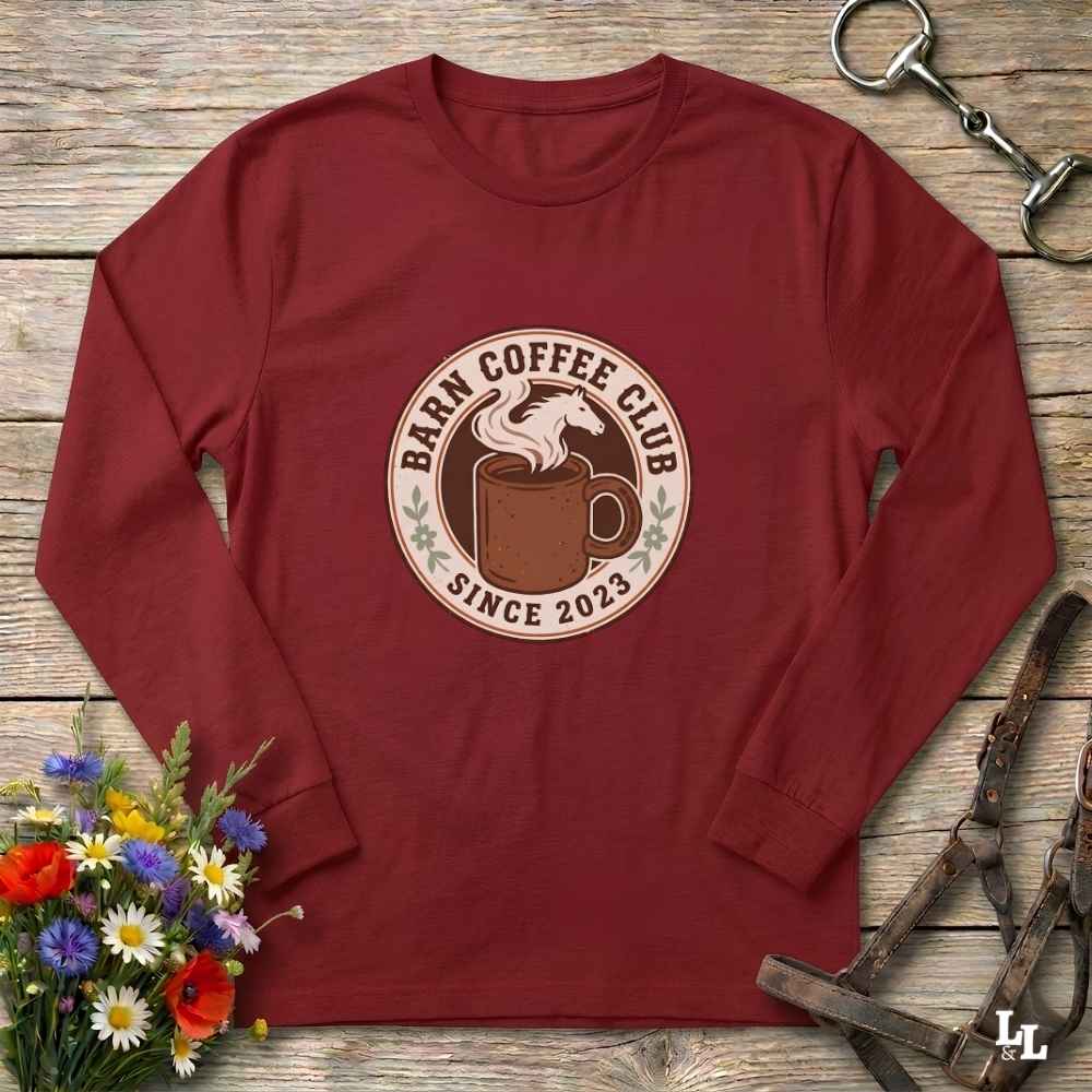 A red long-sleeve t-shirt with a circular 'Barm Coffee Club Since 2023' graphic on the chest, styled in a flat-lay on a rustic wooden background with equestrian and floral props.