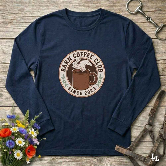 A blue long-sleeve t-shirt with a circular 'Barm Coffee Club Since 2023' graphic on the chest, styled in a flat-lay on a rustic wooden background with equestrian and floral props.