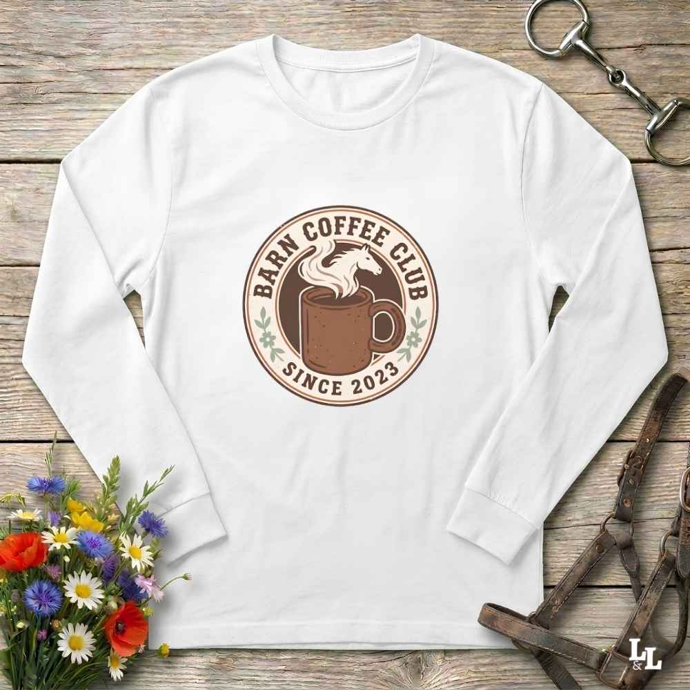 A white long-sleeve t-shirt with a circular 'Barm Coffee Club Since 2023' graphic on the chest, styled in a flat-lay on a rustic wooden background with equestrian and floral props.