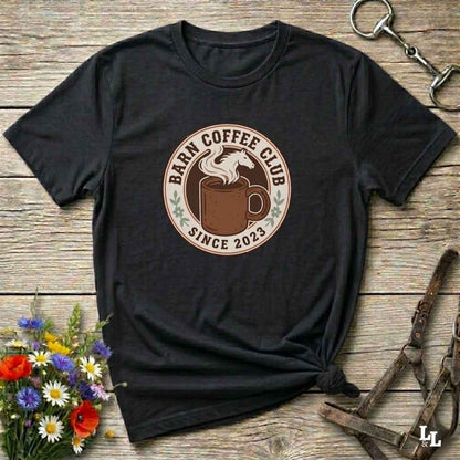 A black short-sleeve graphic T-shirt laid flat on a rustic wooden surface, styled with flowers, a metal key, and leather straps. The shirt has a circular chest print reading “Barn Coffee Club Since 2023” around an illustration of a horse and mug.