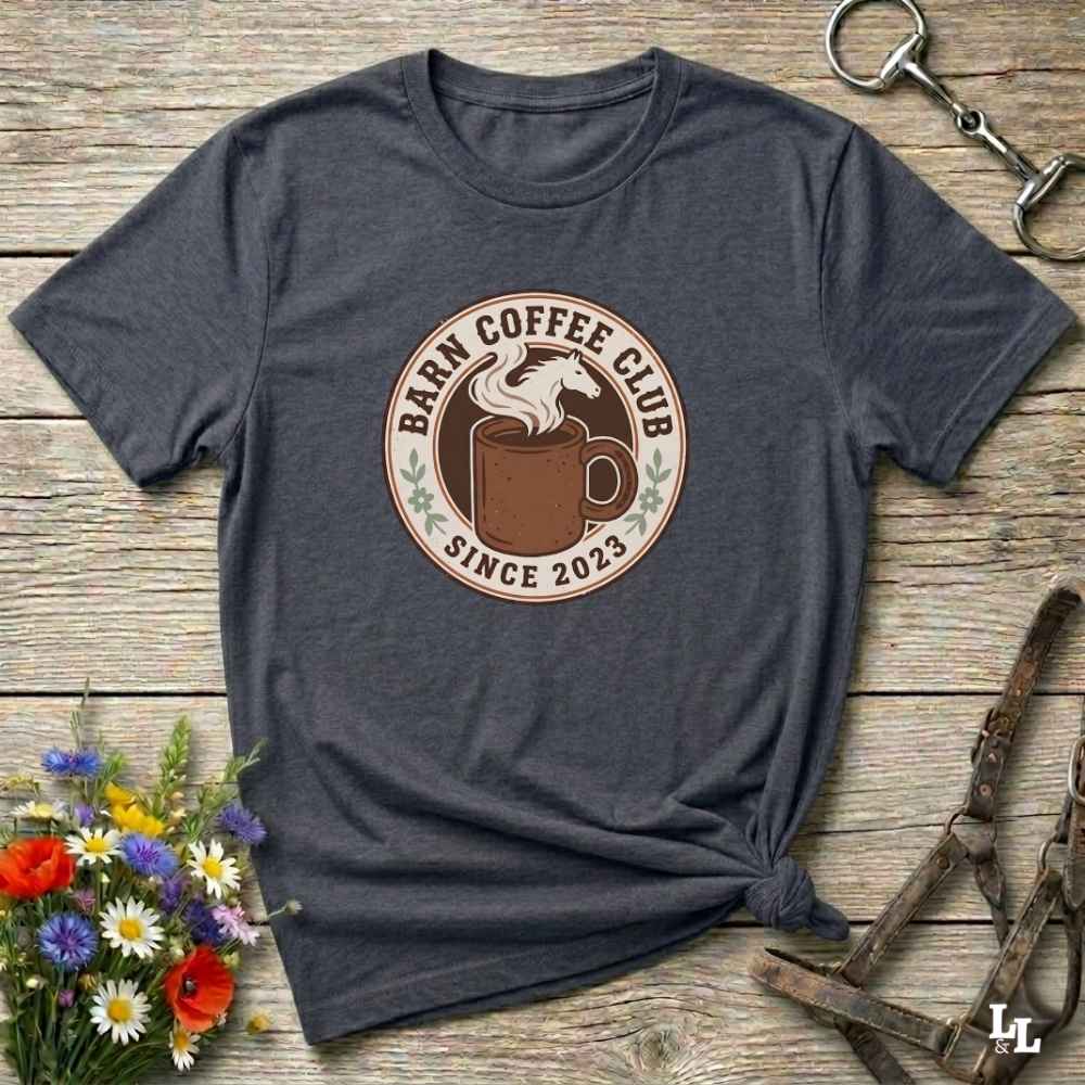 A dark heather gray t-shirt with a circular 'Barn Coffee Club Since 2023' graphic printed on the chest, styled in a flat-lay on a rustic wooden background with a horse halter and flowers.