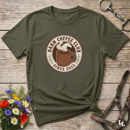 An olive green short-sleeve t-shirt with a circular 'Barn Coffee Club Since 2023' graphic, styled in a flat-lay on a rustic wooden background with a horse bit, leather halter, and a small bouquet of wildflowers.