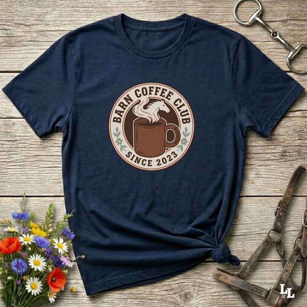 A navy blue short-sleeve t-shirt with a circular 'Barn Coffee Club Since 2023' graphic printed on the chest, styled in a flat-lay on a rustic wooden background with equestrian accessories and flowers.