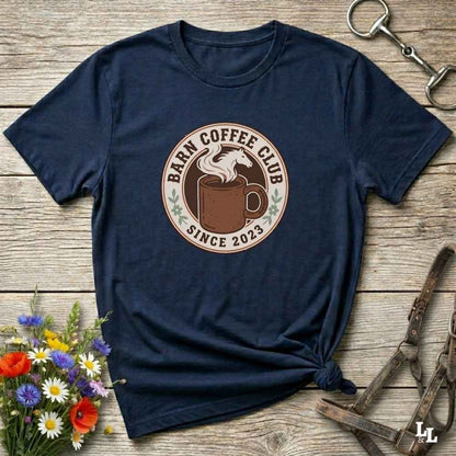A navy blue short-sleeve t-shirt with a circular 'Barn Coffee Club Since 2023' graphic printed on the chest, styled in a flat-lay on a rustic wooden background with equestrian accessories and flowers.