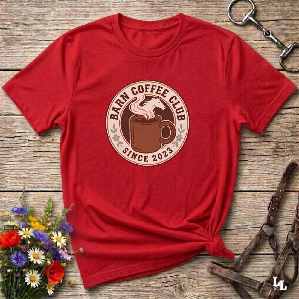 A red short-sleeve t-shirt with a circular 'Barn Coffee Club Since 2023' graphic, styled in a flat-lay on a rustic wooden background with flowers and horse tack.