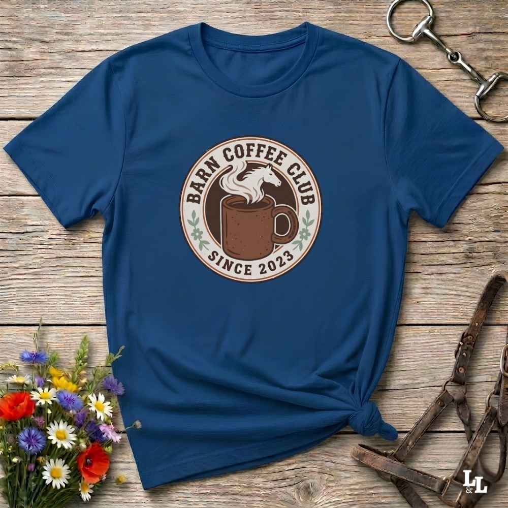 A navy blue short-sleeve t-shirt with a circular 'Barn Coffee Club Since 2023' graphic printed on the chest, styled in a flat-lay on a rustic wooden background with flowers and horse tack.