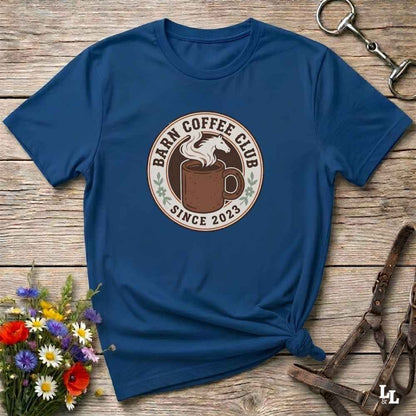 A navy blue short-sleeve t-shirt with a circular 'Barn Coffee Club Since 2023' graphic printed on the chest, styled in a flat-lay on a rustic wooden background with flowers and horse tack.