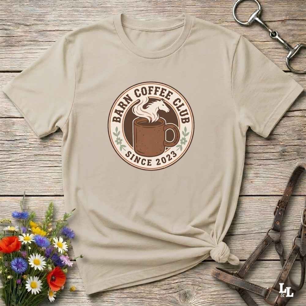 A beige short-sleeve t-shirt with a circular 'Barn Coffee Club' graphic, styled in a flat-lay on a rustic wooden background with flowers and leather tack.