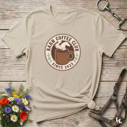 A beige short-sleeve t-shirt with a circular 'Barn Coffee Club' graphic, styled in a flat-lay on a rustic wooden background with flowers and leather tack.