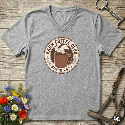 A heather gray V-neck t-shirt laid flat on a rustic wooden surface, featuring a circular 'Farm Coffee Club Since 2023' graphic on the chest with a coffee mug and horse illustration. The shirt is styled with equestrian tack and a small bouquet of wildflowers.