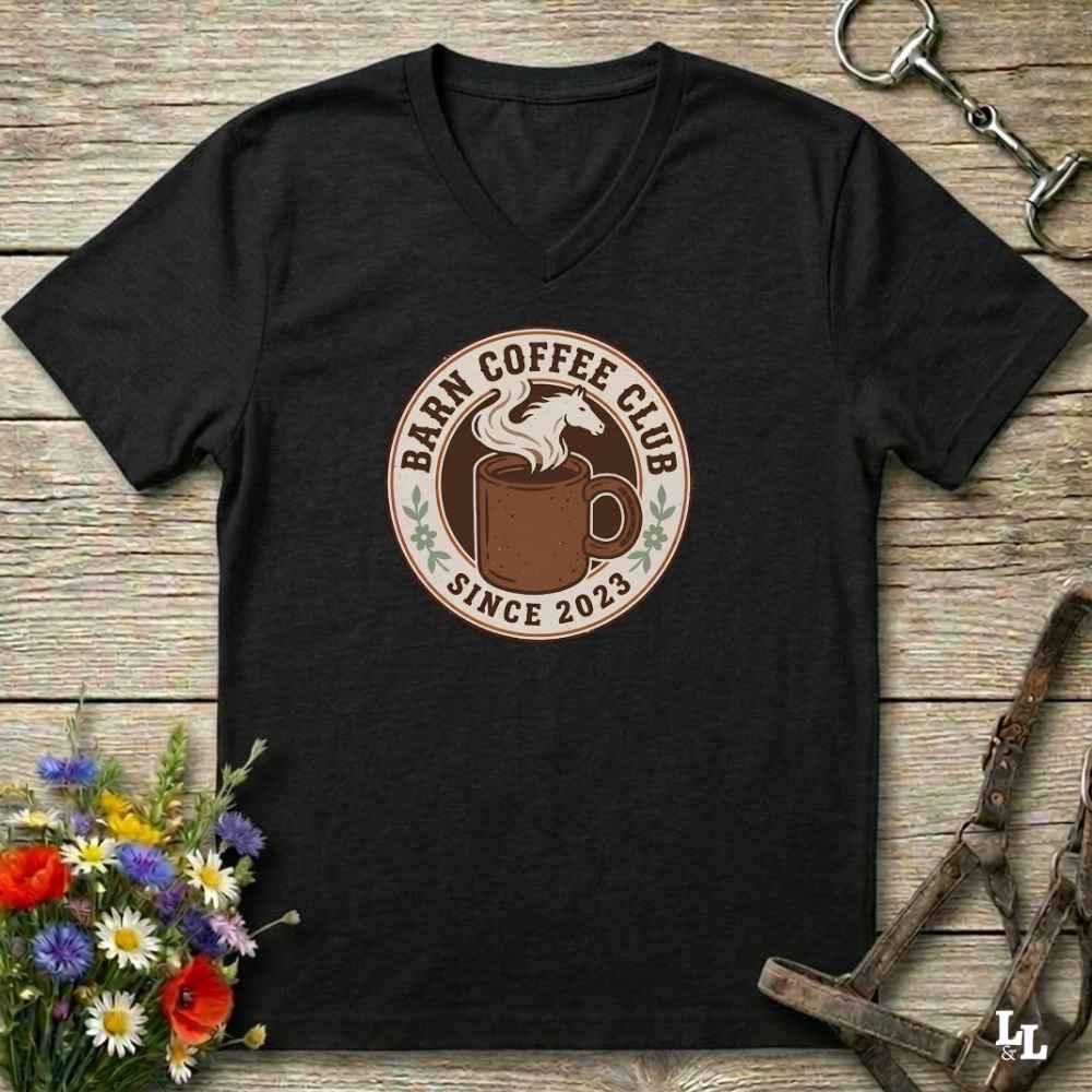 A black V-neck t-shirt with a circular 'Farm Coffee Club Since 2023' graphic on the chest, displayed flat on a rustic wooden background with equestrian accessories and flowers.