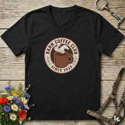 A black V-neck t-shirt with a circular 'Farm Coffee Club Since 2023' graphic on the chest, displayed flat on a rustic wooden background with equestrian accessories and flowers.