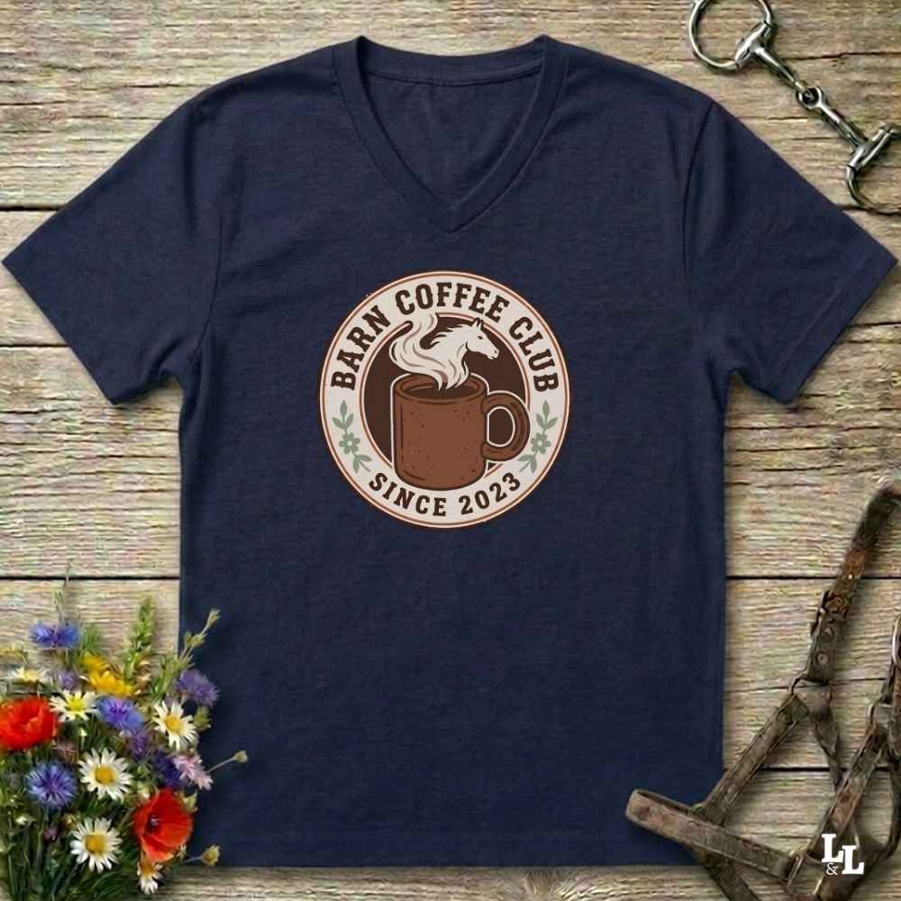 A navy blue v-neck t-shirt with a circular 'Barm Coffee Club Since 2023' graphic on the chest, displayed in a flat-lay arrangement on a rustic wooden background with equestrian accessories and flowers.
