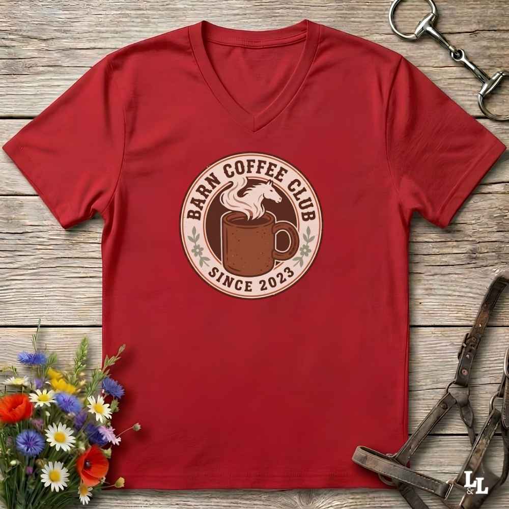 A red v-neck t-shirt with a circular 'Barm Coffee Club Since 2023' graphic on the chest, displayed in a flat-lay arrangement on a rustic wooden background with equestrian accessories and flowers.